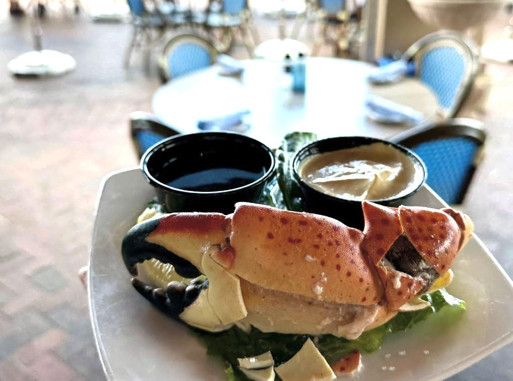 A close-up of a chilled stone crab claw served on lettuce with lemon wedges and small cups of melted butter and mustard sauce, set against a softly blurred patio dining area.