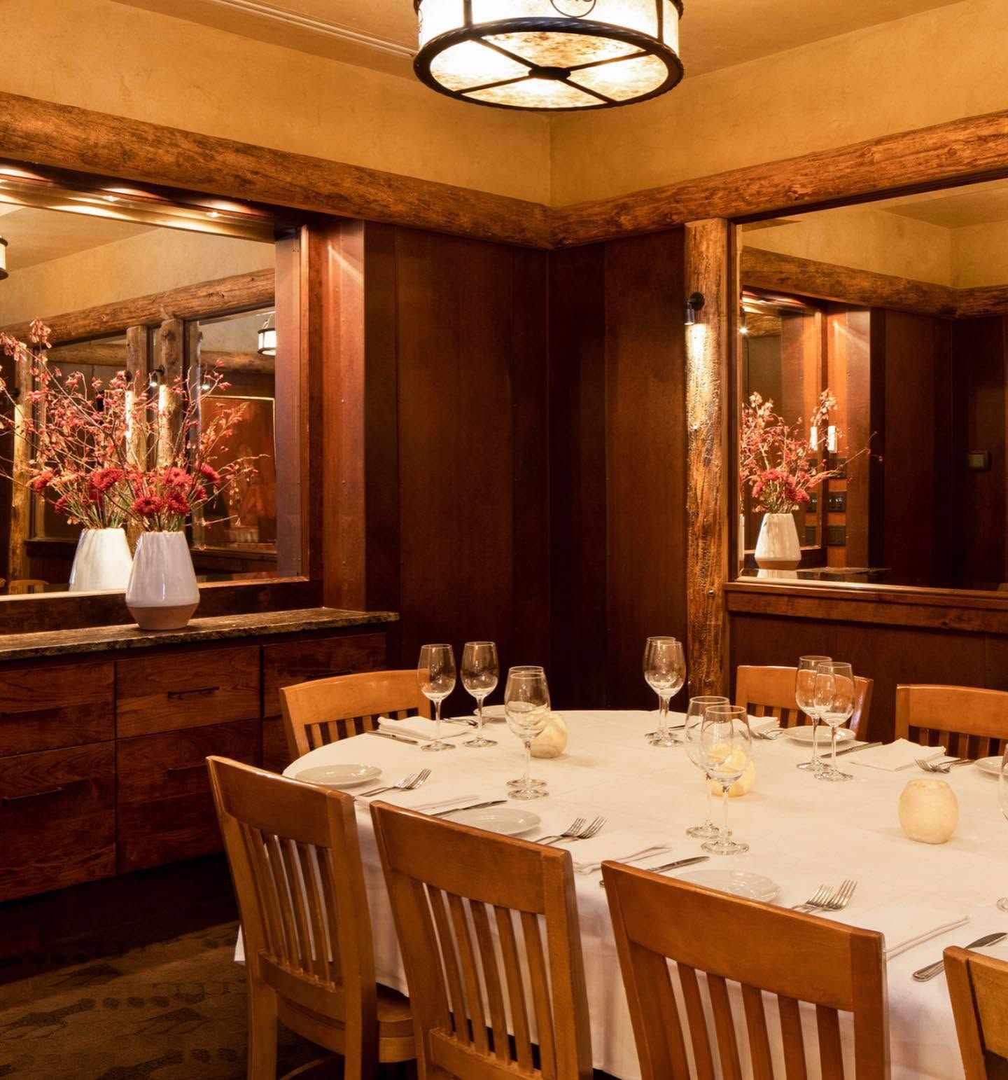 A round table set with crisp white linens, polished wine glasses, and candlelight anchors an intimate dining room lined with rich wood paneling and exposed log beams. Mirrored walls reflect vases of coral-toned flowers, creating a warm, upscale mountain-lodge atmosphere. 