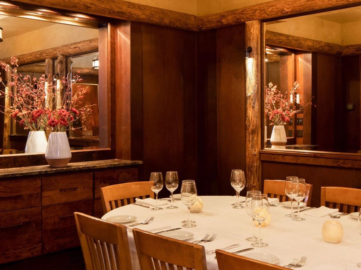 A round table set with crisp white linens, polished wine glasses, and candlelight anchors an intimate dining room lined with rich wood paneling and exposed log beams. Mirrored walls reflect vases of coral-toned flowers, creating a warm, upscale mountain-lodge atmosphere.
