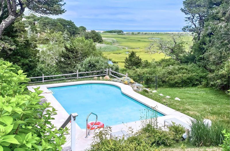 Saltwater Serenity features a private outdoor pool overlooking green coastal marshland with the ocean visible in the distance. The quiet setting gives guests a peaceful place to swim and enjoy the natural scenery of Cape Cod.