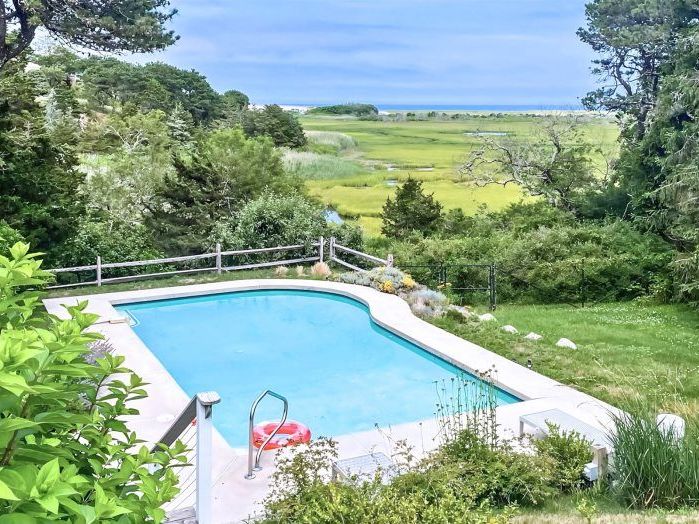 Saltwater Serenity features a private outdoor pool overlooking green coastal marshland with the ocean visible in the distance. The quiet setting gives guests a peaceful place to swim and enjoy the natural scenery of Cape Cod.