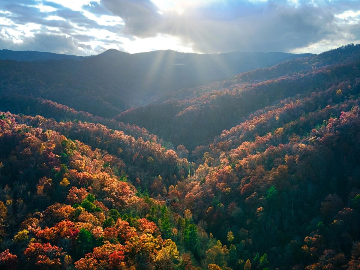 Sun rays break through clouds over a valley of forested mountains, where autumn foliage glows in shades of red, orange, and gold.