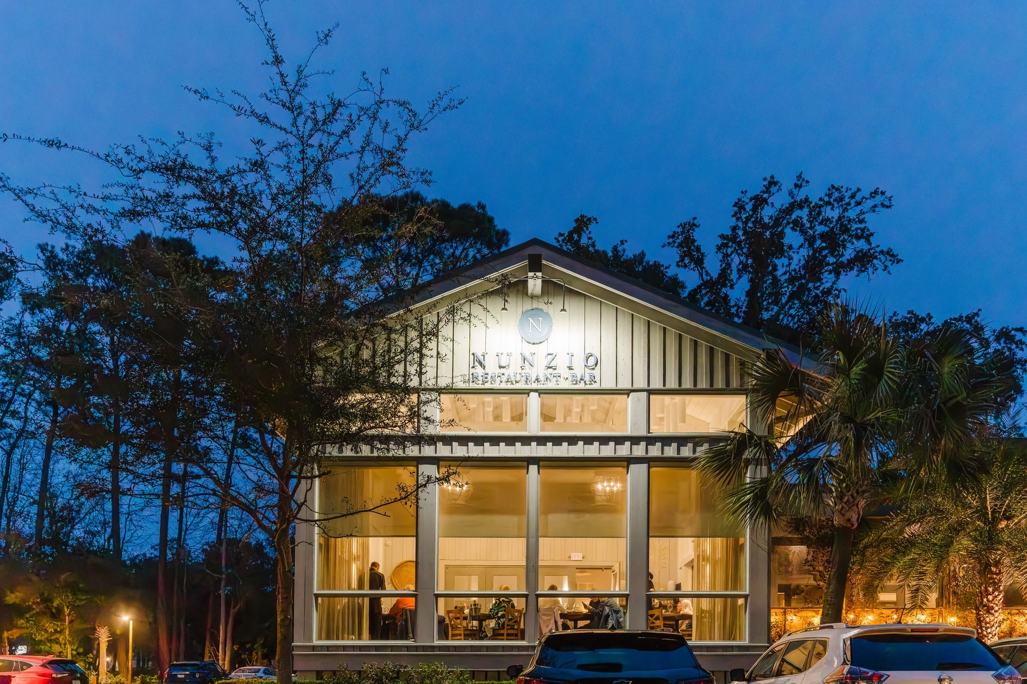 NUNZIO Restaurant + Bar glowing at night with warm lights shining through tall windows on Hilton Head Island. Guests dine inside the modern coastal space surrounded by palm trees and quiet evening skies. This upscale Hilton Head restaurant is known for refined Italian dishes and an intimate dining atmosphere.