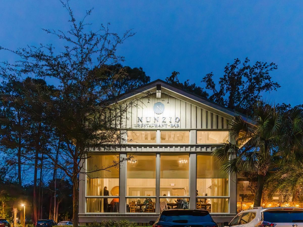 NUNZIO Restaurant + Bar glowing at night with warm lights shining through tall windows on Hilton Head Island. Guests dine inside the modern coastal space surrounded by palm trees and quiet evening skies. This upscale Hilton Head restaurant is known for refined Italian dishes and an intimate dining atmosphere.
