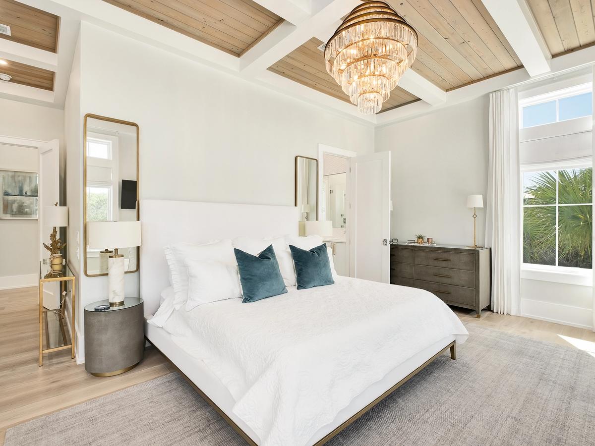 A bright and elegant bedroom featuring a white bed with teal accent pillows, gold and marble decor, a coffered wood ceiling, and a sparkling chandelier above.