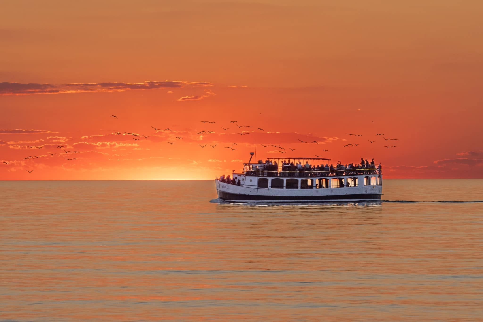 A classic double-decker tour boat glides across calm water as the sun sinks into a fiery orange horizon, with a flock of birds scattered across the glowing sky. Passengers fill the open upper deck, soaking in the vivid Lake Michigan sunset. 