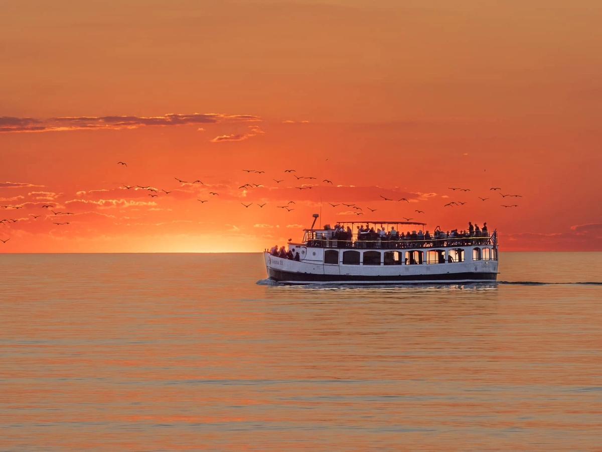 A classic double-decker tour boat glides across calm water as the sun sinks into a fiery orange horizon, with a flock of birds scattered across the glowing sky. Passengers fill the open upper deck, soaking in the vivid Lake Michigan sunset.