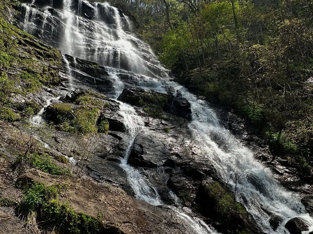 A stunning waterfall cascades down rugged rock surrounded by lush green trees. Sunlight sparkles on the water as it tumbles down, creating a peaceful mountain scene perfect for hikers and nature lovers.