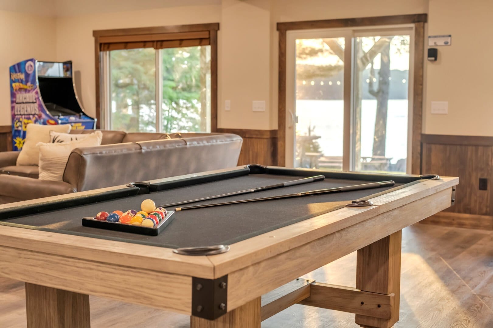 A rustic game room featuring a wooden pool table with racked billiard balls and cues, set against warm wood paneling, a leather sofa, arcade machine, and sliding glass doors overlooking a wooded outdoor area.