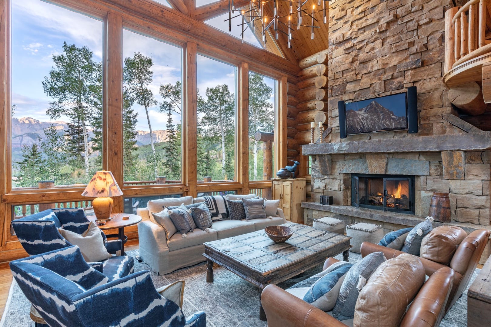 Cozy mountain living room with large windows, stone fireplace, rustic wood beams, and comfortable seating overlooking forested peaks.