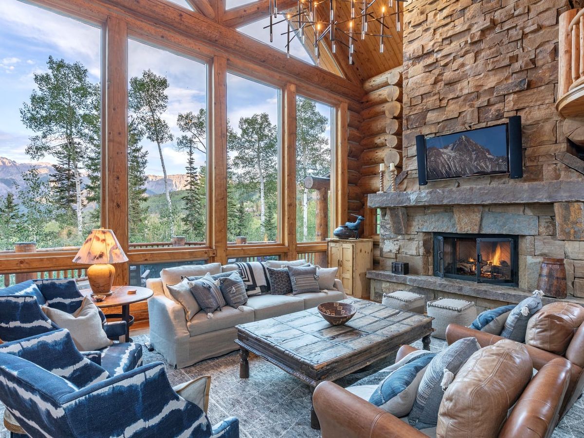 Cozy mountain living room with large windows, stone fireplace, rustic wood beams, and comfortable seating overlooking forested peaks.