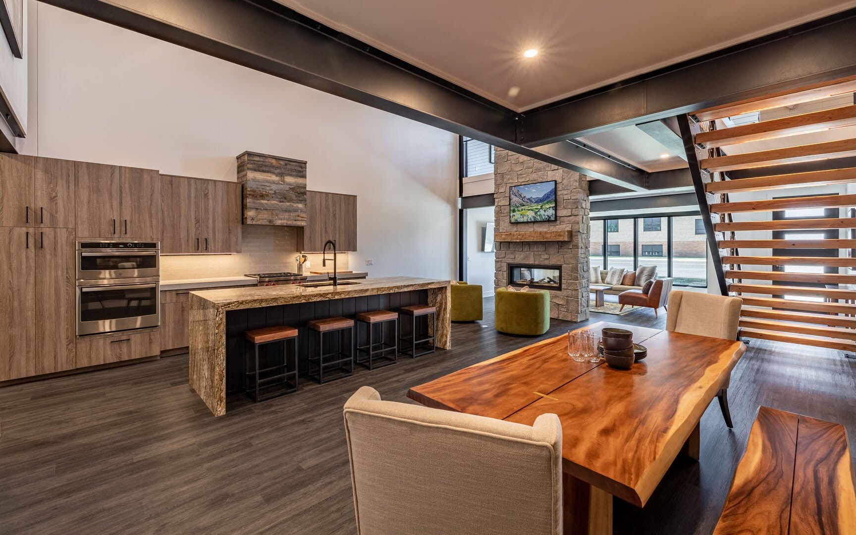 Modern open-concept kitchen and living area featuring wood cabinetry, a stone fireplace, a large natural wood dining table, and a floating staircase with horizontal wooden slats.