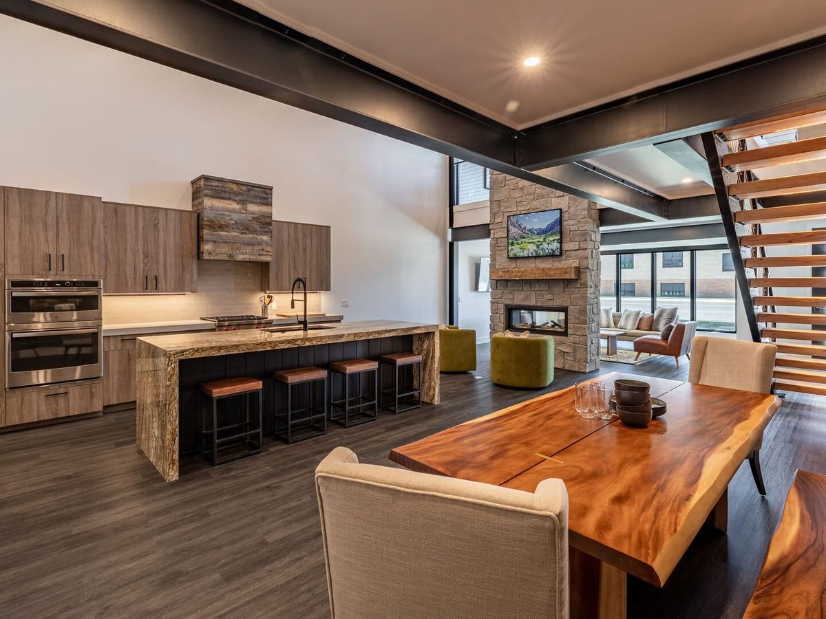 Modern open-concept kitchen and living area featuring wood cabinetry, a stone fireplace, a large natural wood dining table, and a floating staircase with horizontal wooden slats.