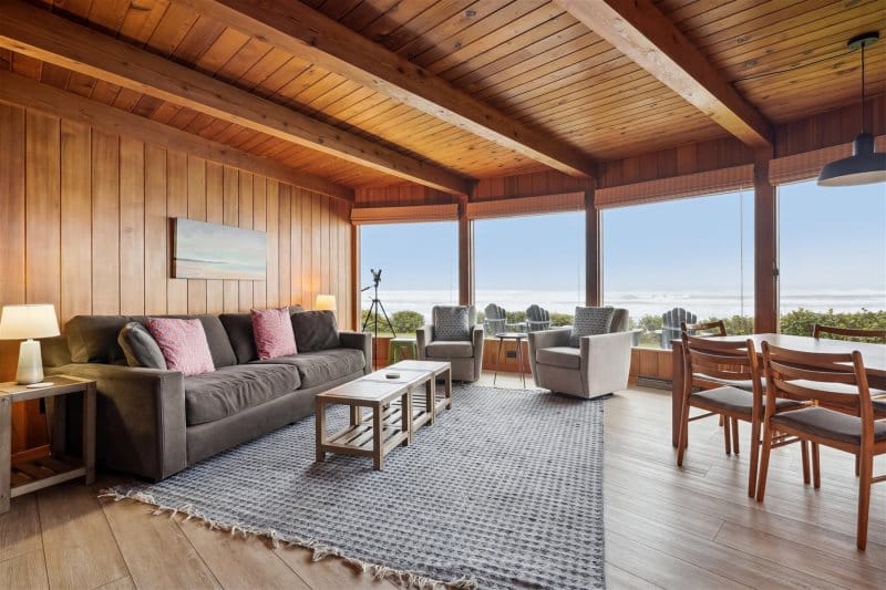 A cozy wood-paneled living room with ocean views through large windows, offering a warm, rustic charm. Perfect for relaxing with family, it brings together coastal beauty and cabin-style comfort.