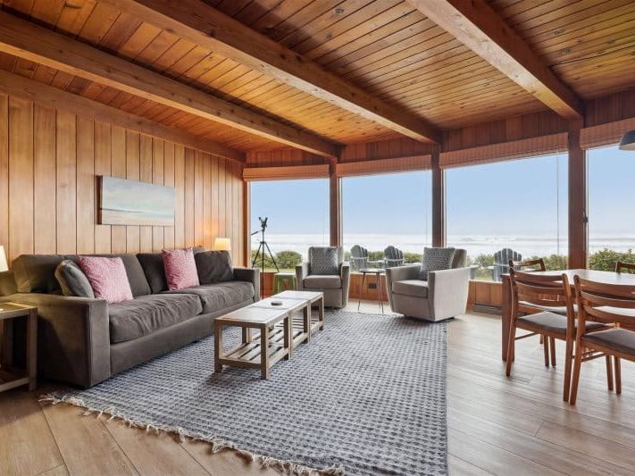 A cozy wood-paneled living room with ocean views through large windows, offering a warm, rustic charm. Perfect for relaxing with family, it brings together coastal beauty and cabin-style comfort.