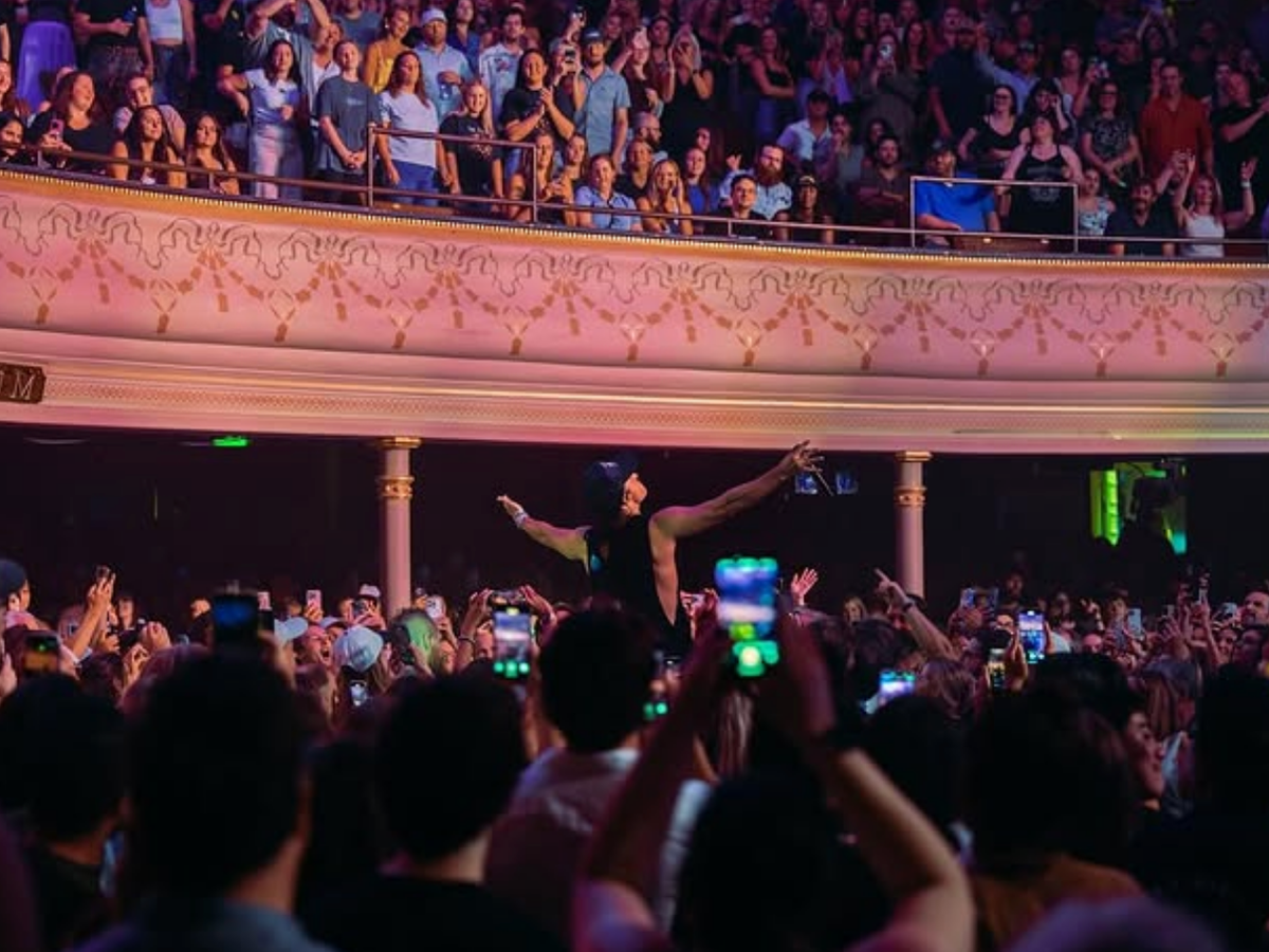 A lively concert scene with a performer standing in the crowd, arms outstretched, as fans capture the moment on their phones inside a packed theater.