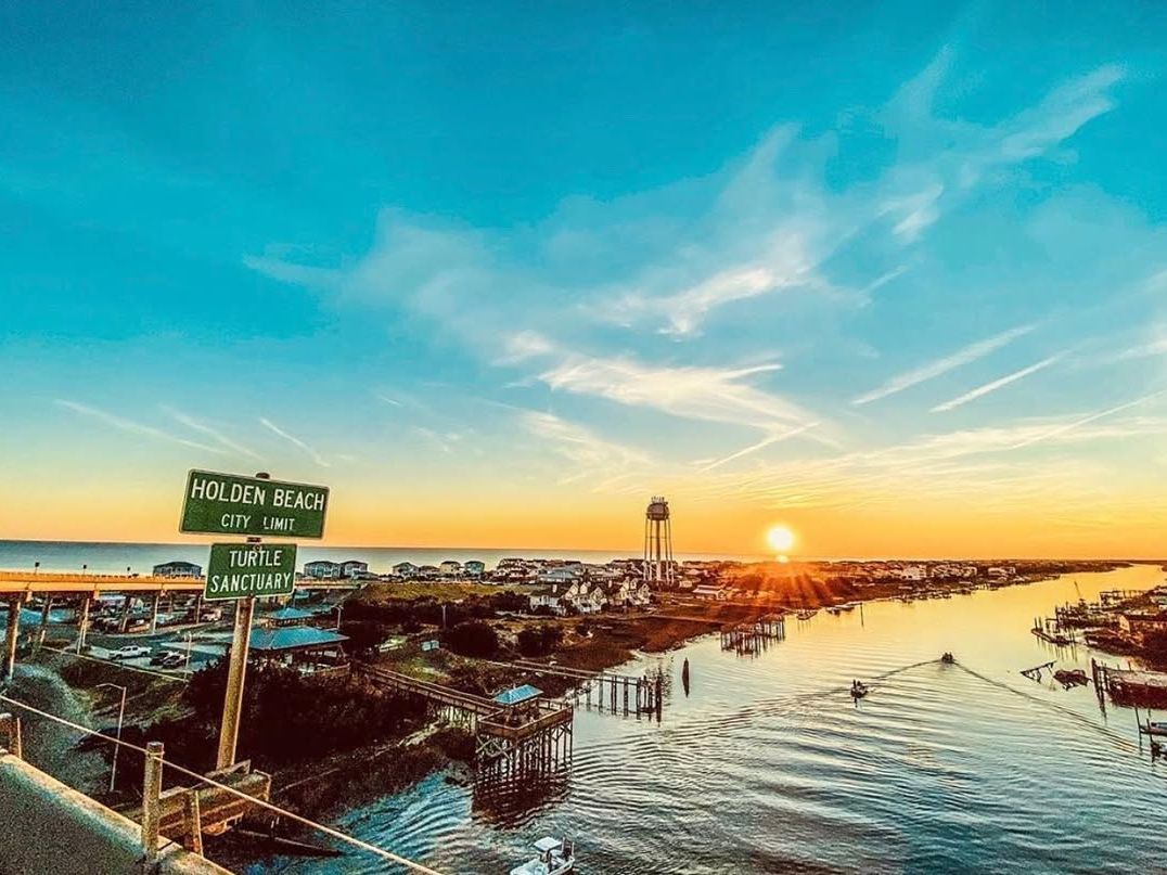 A sunset sky glows above Holden Beach with the city sign and turtle sanctuary marker in view. Boats cruise the inlet below a colorful sky.