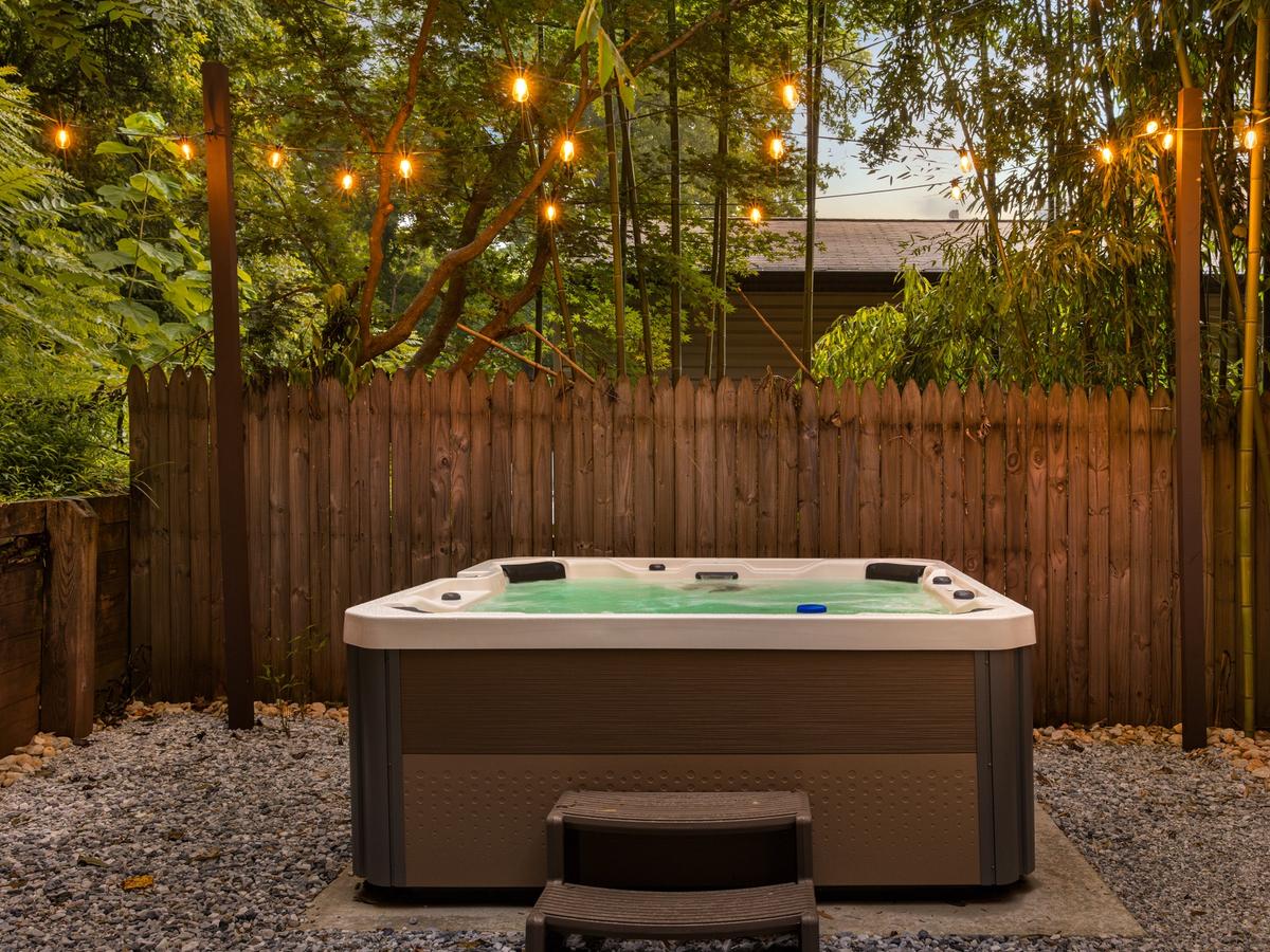 A cozy backyard hot tub setup surrounded by a wooden privacy fence and lush greenery, with string lights softly glowing above on a gravel-covered patio.