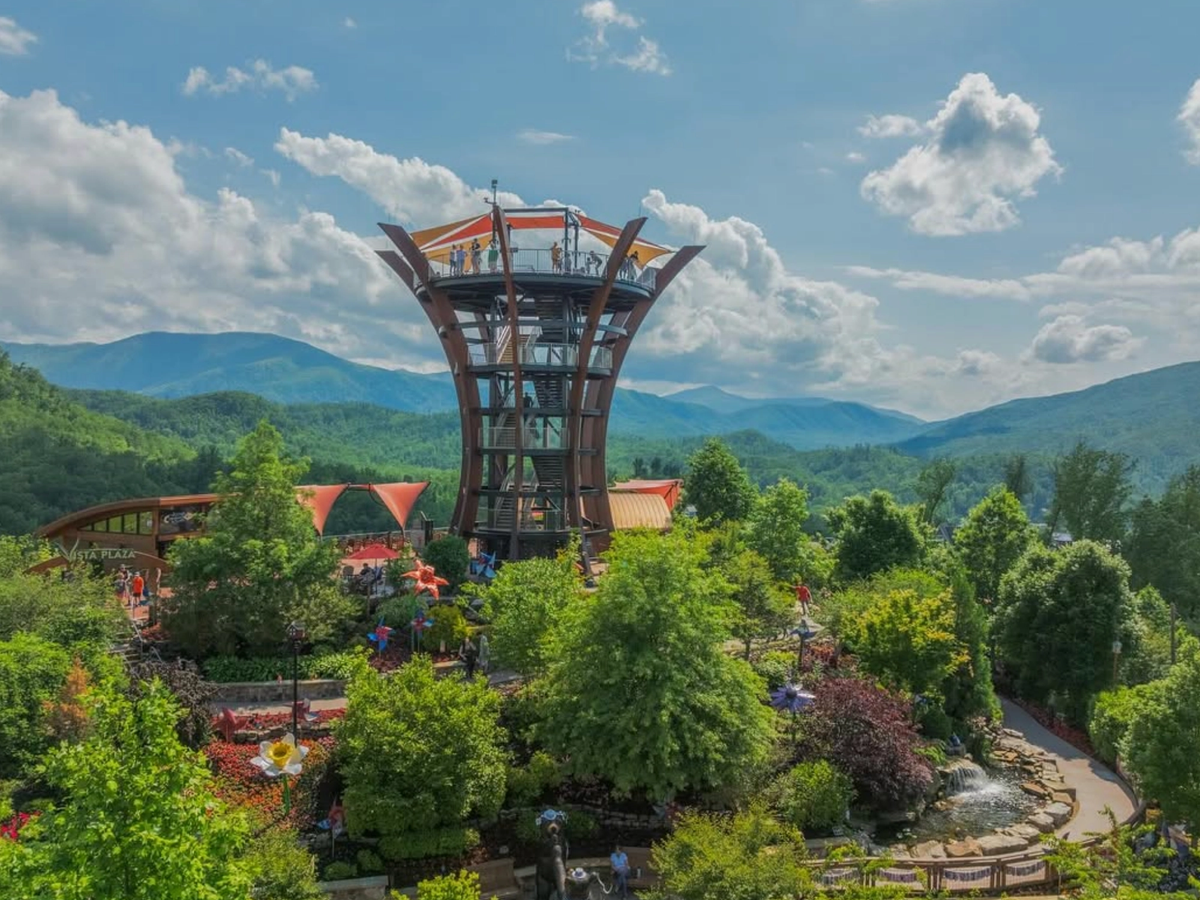A scenic observation tower surrounded by lush gardens and trees, set against a backdrop of rolling Smoky Mountain peaks under a partly cloudy sky.