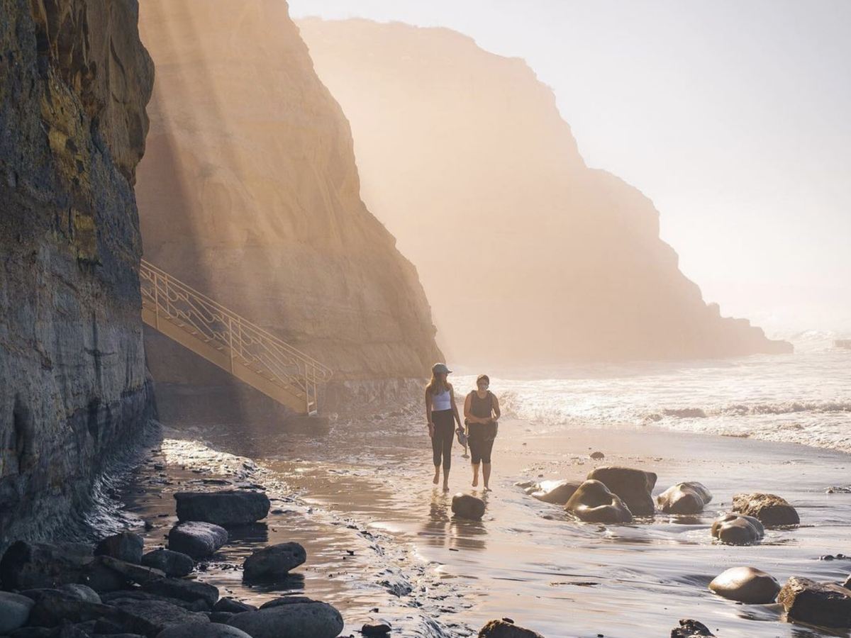 Two people walk along a rocky beach at the base of tall cliffs, with golden sunlight streaming through the mist and waves rolling in from the ocean.