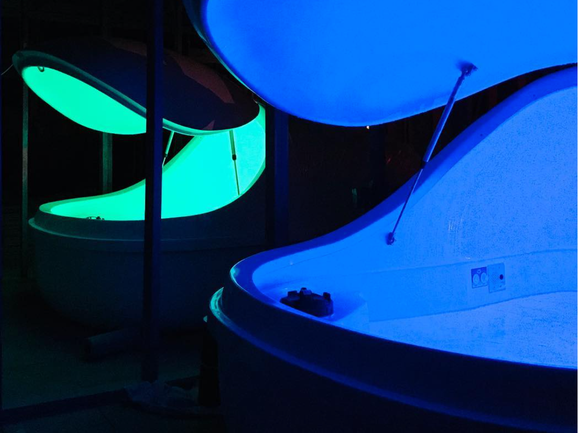 Two sensory deprivation float pods glow with calming green and blue light in a dark, tranquil spa setting.