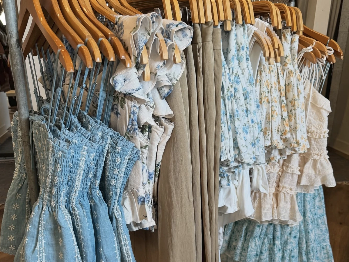 A clothing rack displaying a collection of light and airy women’s summer tops and dresses in soft pastel colors, featuring floral prints, ruffles, and smocked detailing.