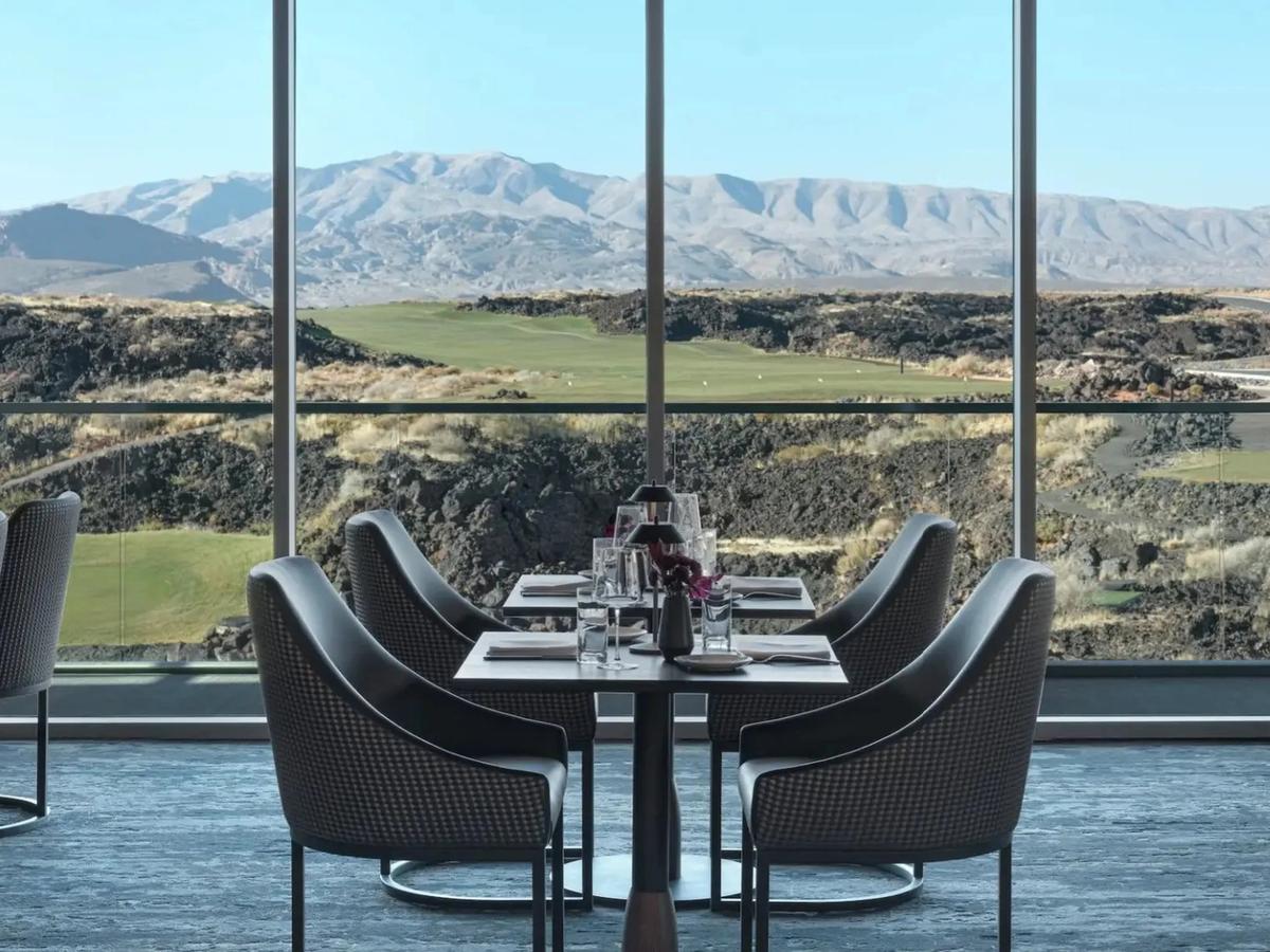 A sleek, upscale restaurant with floor-to-ceiling windows framing a scenic desert mountain golf course view. Elegant table settings and plush seating create a refined dining experience in the heart of the southwest.