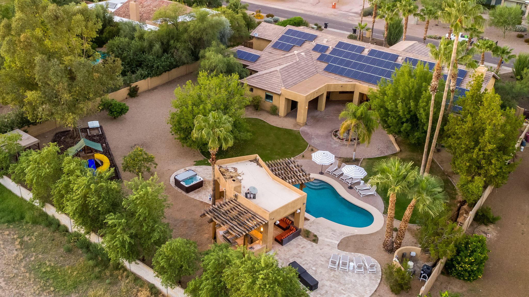 An aerial view of a desert property with a pool, hot tub, and lounge chairs, surrounded by trees and solar panels on the roof. A play area with a slide is also visible in the yard.