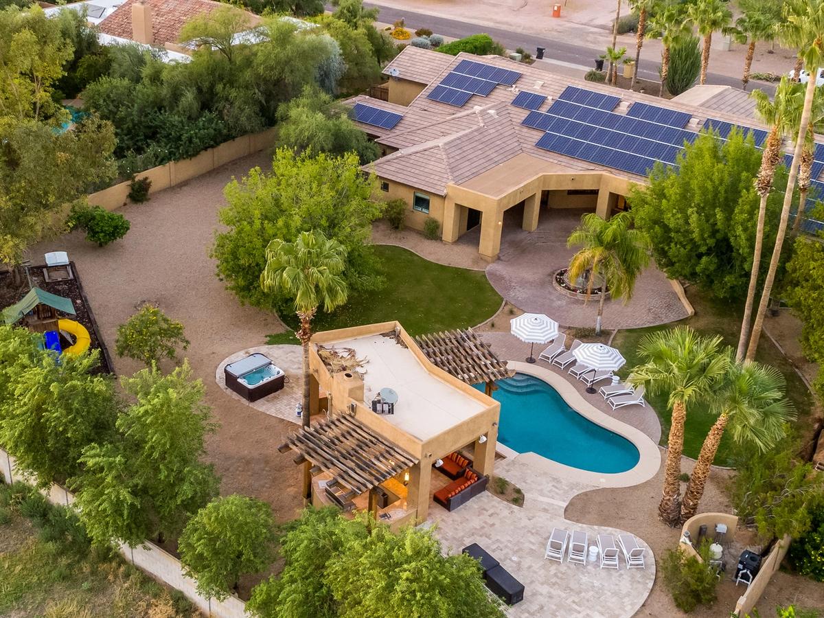 An aerial view of a desert property with a pool, hot tub, and lounge chairs, surrounded by trees and solar panels on the roof. A play area with a slide is also visible in the yard.