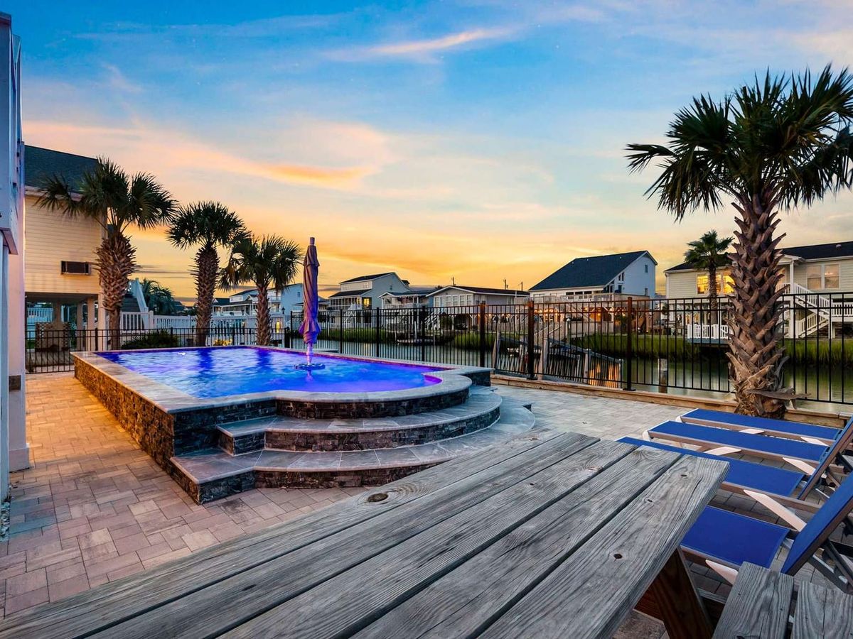 A glowing infinity pool with purple lighting is surrounded by palm trees, lounge chairs, and a canal backdrop at sunset.