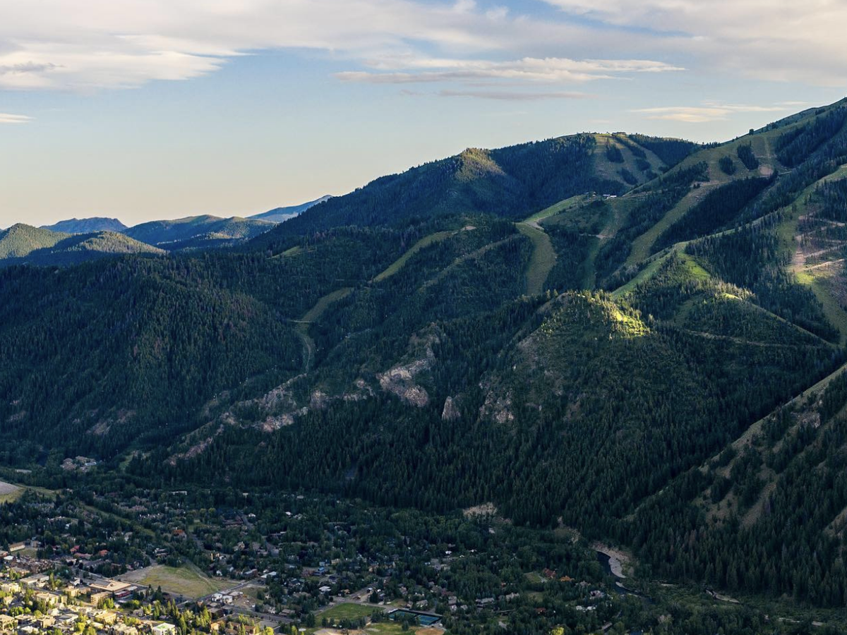 Aerial view of a lush, forested mountain range with ski slopes and a small town nestled in the valley below under a partly cloudy sky.