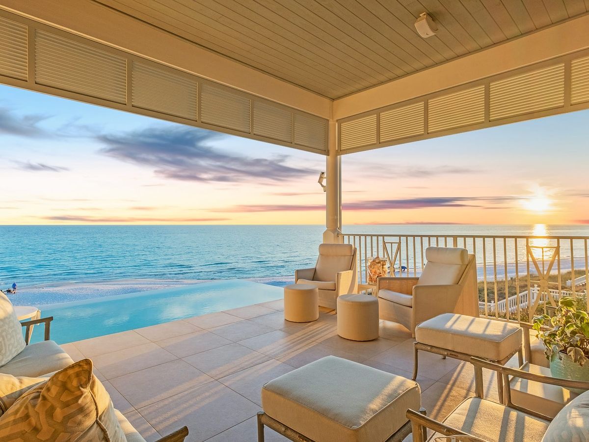 : A stylish covered terrace with plush seating and an infinity pool facing the sunset over the Gulf of Mexico. This dreamy coastal view captures the beauty of 30A evenings in pure luxury.