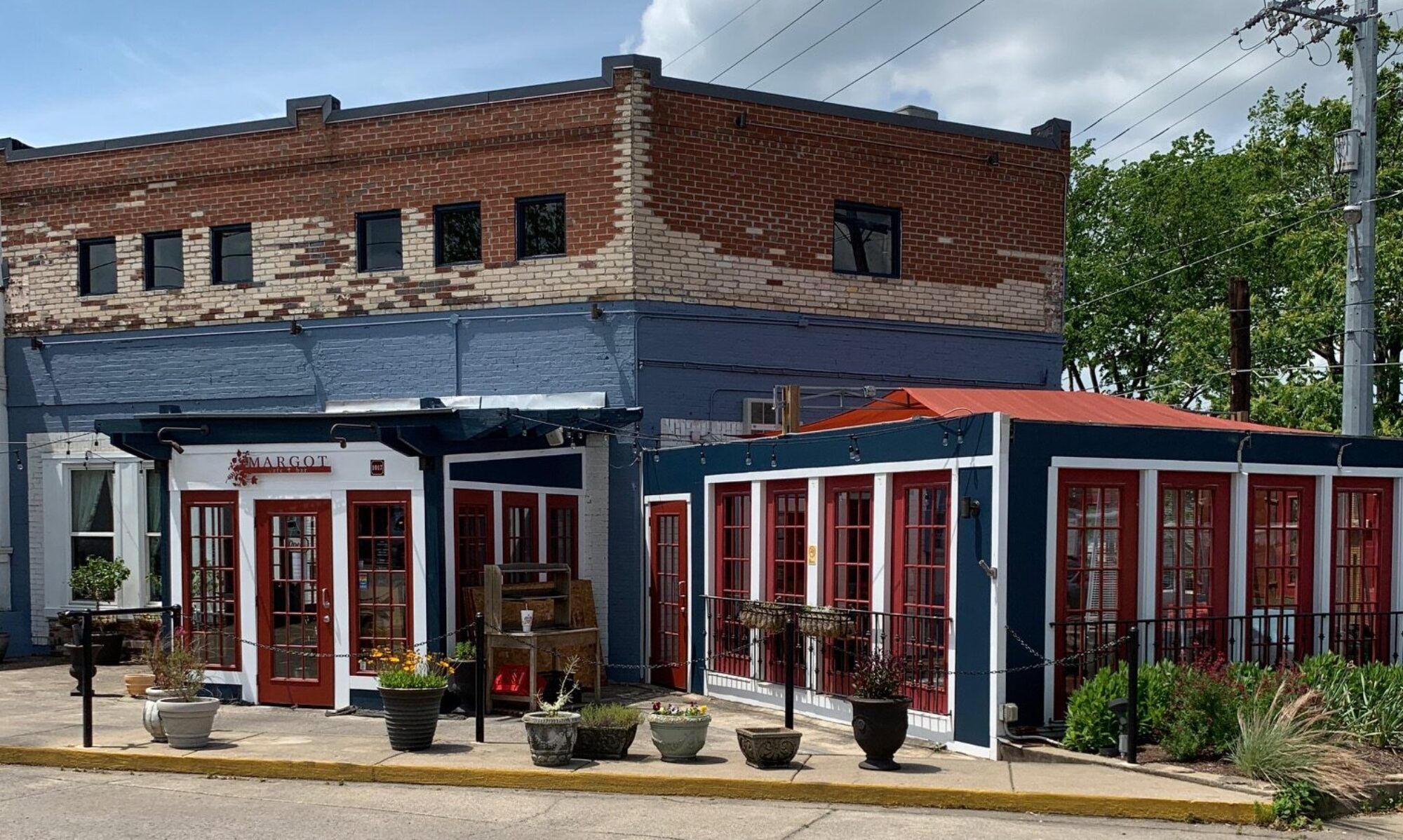 A quaint European-style restaurant with red-framed windows and a rustic brick exterior. Potted plants and outdoor seating create a welcoming neighborhood charm perfect for a relaxed meal.