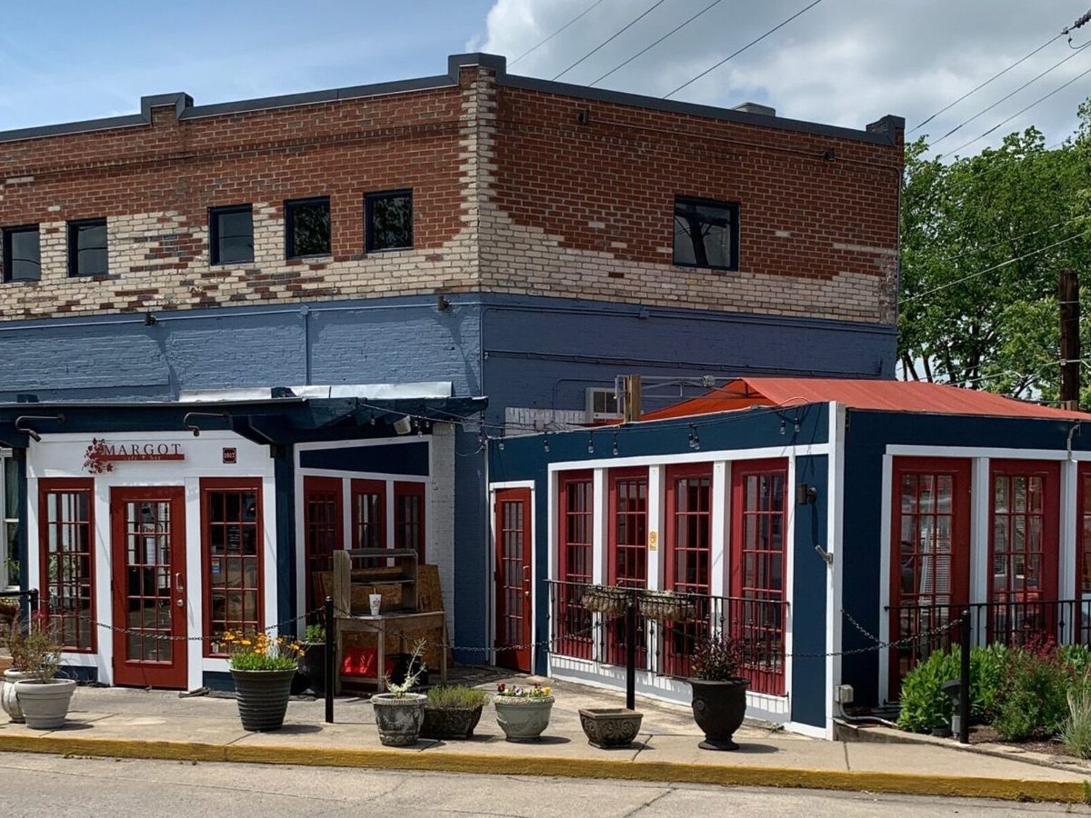 A quaint European-style restaurant with red-framed windows and a rustic brick exterior. Potted plants and outdoor seating create a welcoming neighborhood charm perfect for a relaxed meal.