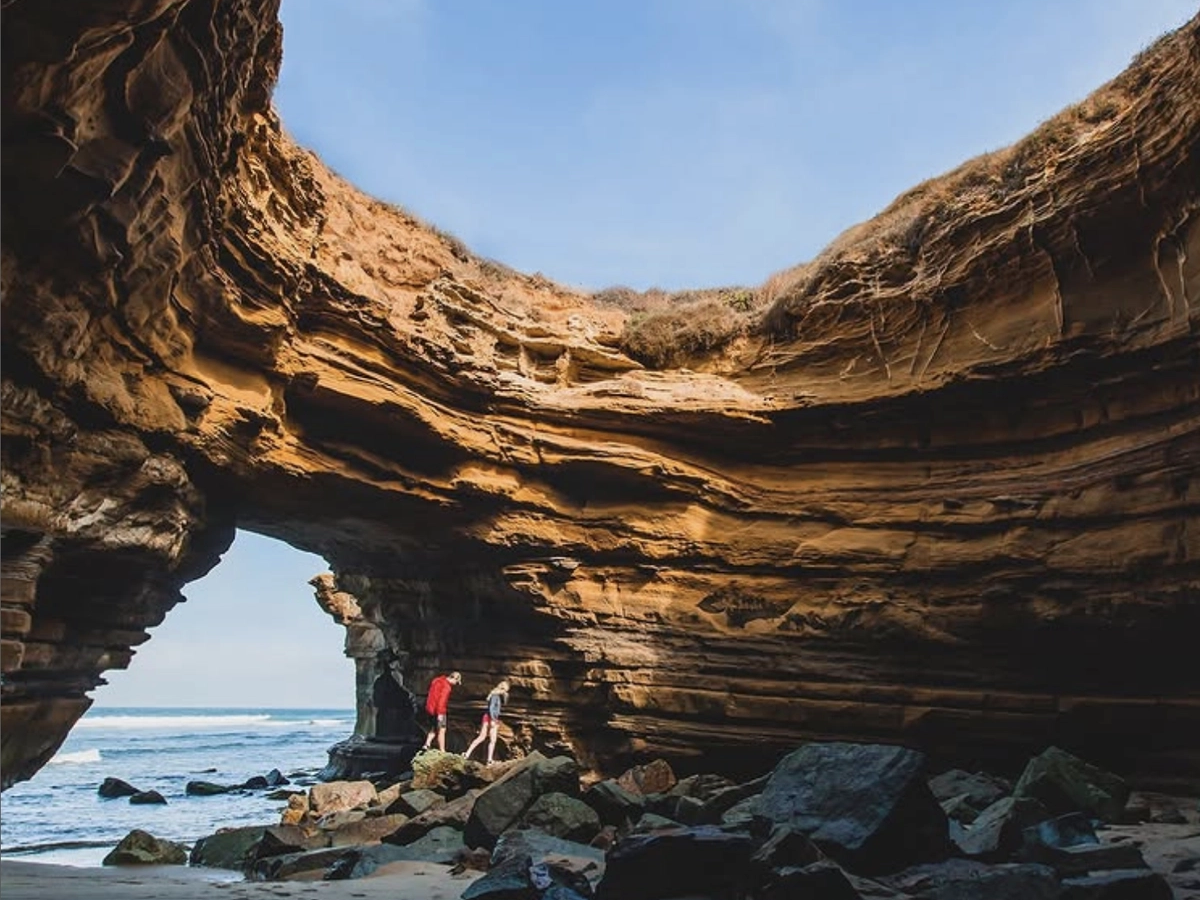 Two people explore inside a dramatic sea cave with layered sandstone walls, a rocky floor, and an opening that reveals blue sky and the ocean beyond.