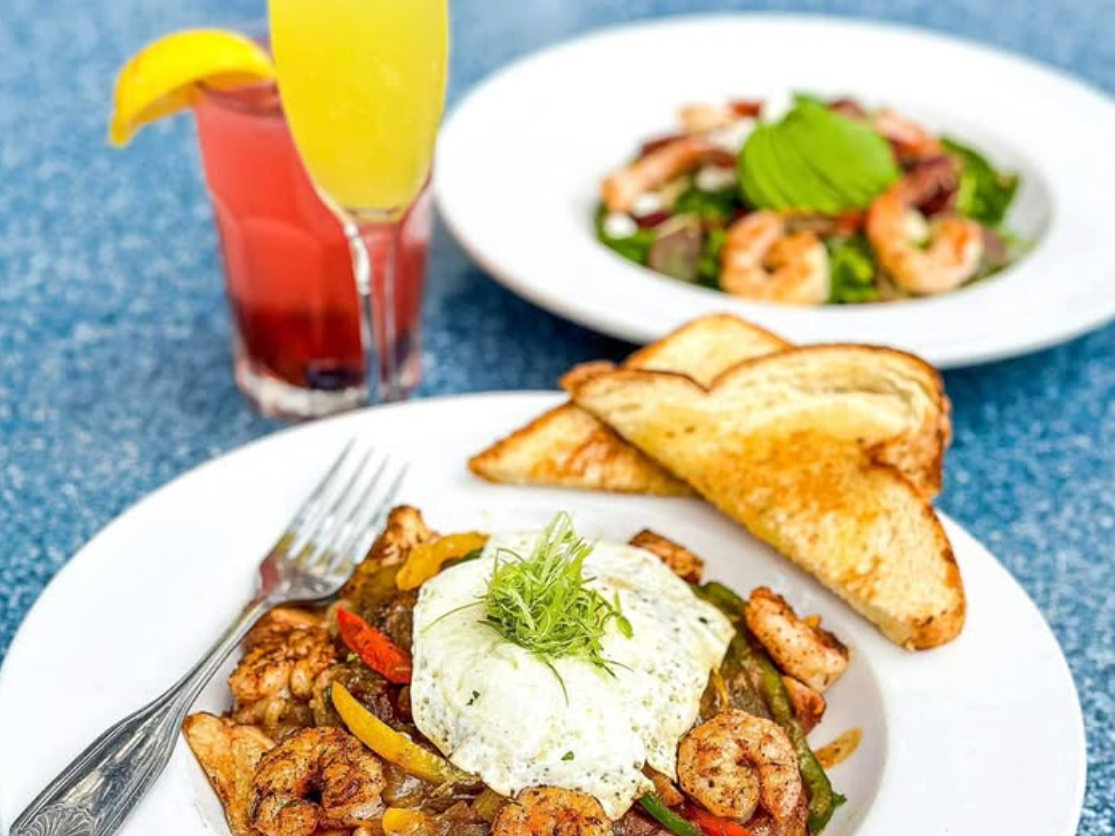 A brunch spread with a plate of shrimp, roasted potatoes, peppers, and a fried egg on top, served with toast; behind it sits a shrimp salad and two cocktails, one garnished with an orange slice.