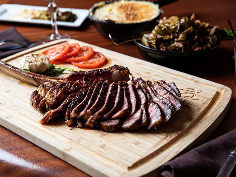 Juicy tomahawk steak sliced and served on a wooden board with grilled garlic and tomatoes. A mouthwatering choice for meat lovers in Charleston.