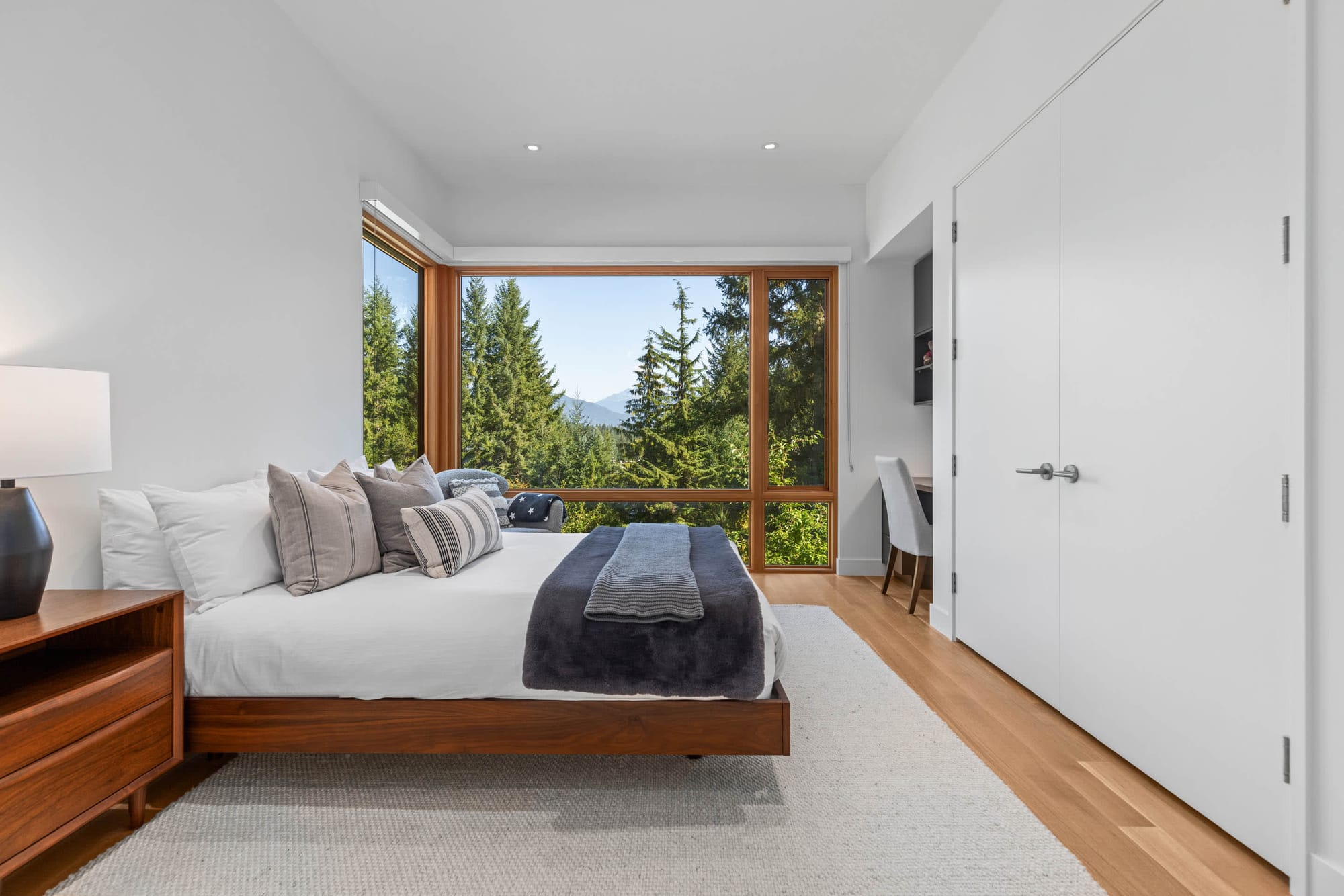 Bright, minimalist bedroom with a low wood platform bed, neutral linens, and a floor-to-ceiling picture window overlooking lush evergreen trees and distant mountains.