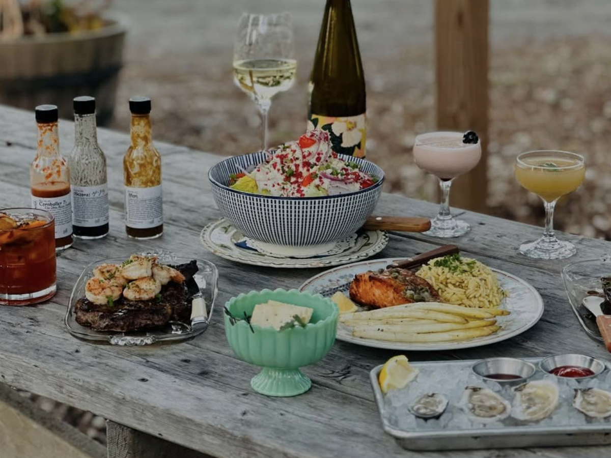 A rustic outdoor dining setup featuring gourmet dishes, including steak with shrimp, salmon with orzo and asparagus, oysters, and a fresh salad. The table is adorned with craft cocktails, wine, and bottles of artisanal hot sauce, creating a cozy and inviting atmosphere