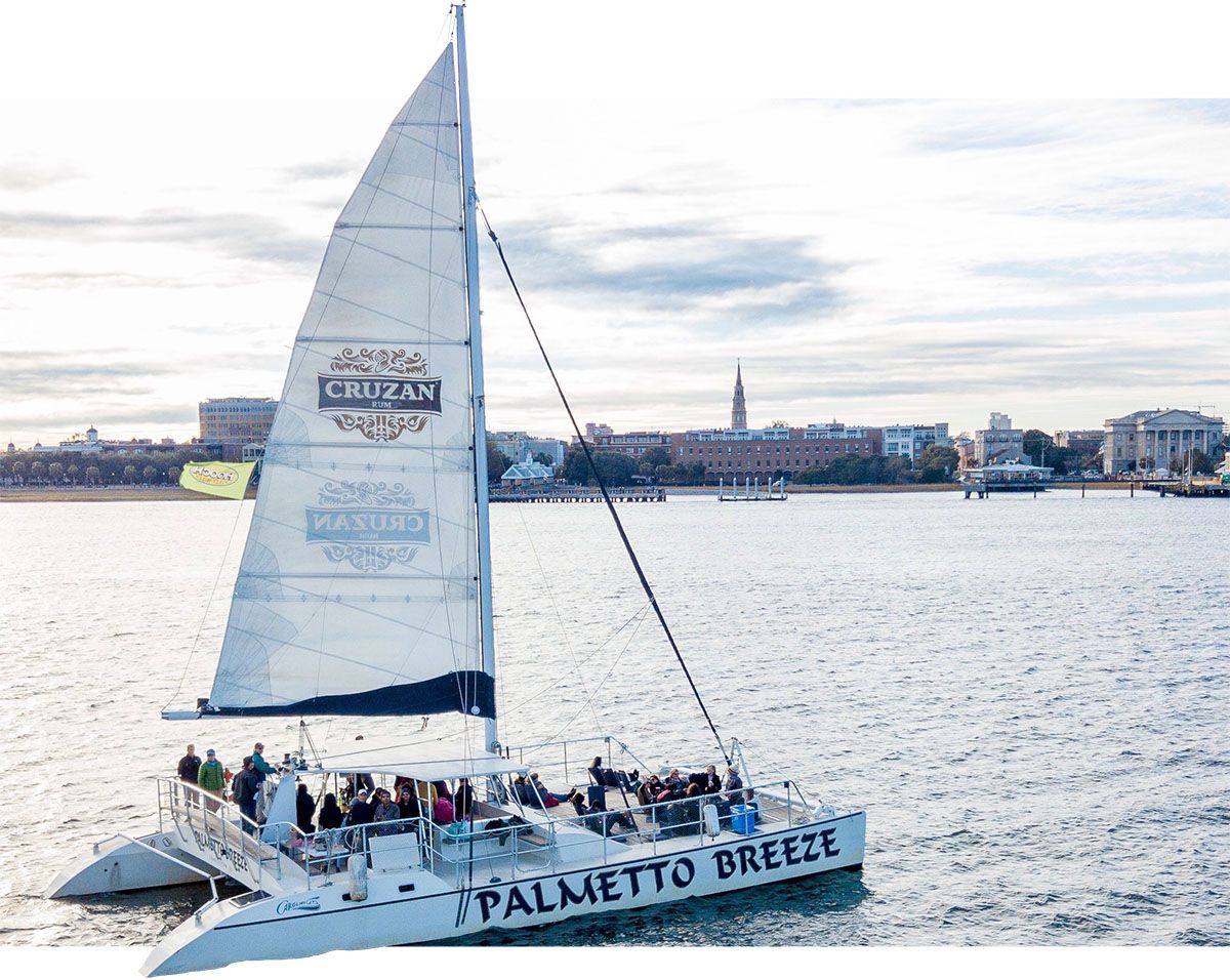 The Palmetto Breeze catamaran sails through Charleston Harbor with passengers enjoying the views. Its tall white sail and spacious deck make it a favorite for sunset cruises and private charters