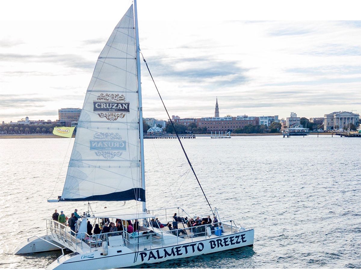 The Palmetto Breeze catamaran sails through Charleston Harbor with passengers enjoying the views. Its tall white sail and spacious deck make it a favorite for sunset cruises and private charters