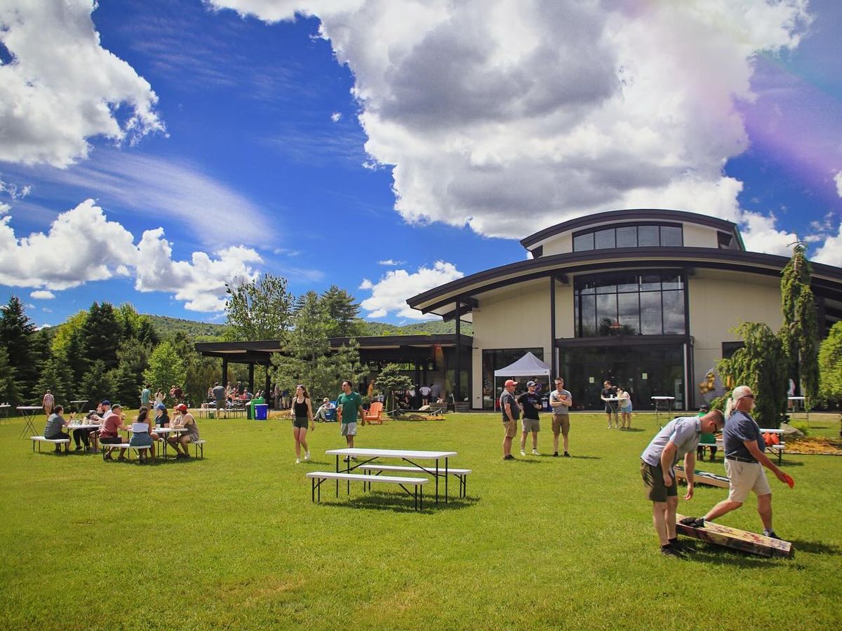 Locals and travelers gather on a sunny afternoon at The Alchemist Brewery, enjoying craft beer and games on the grassy lawn. The modern brewery’s glass architecture gleams under a vibrant blue sky as laughter and conversation fill the air.