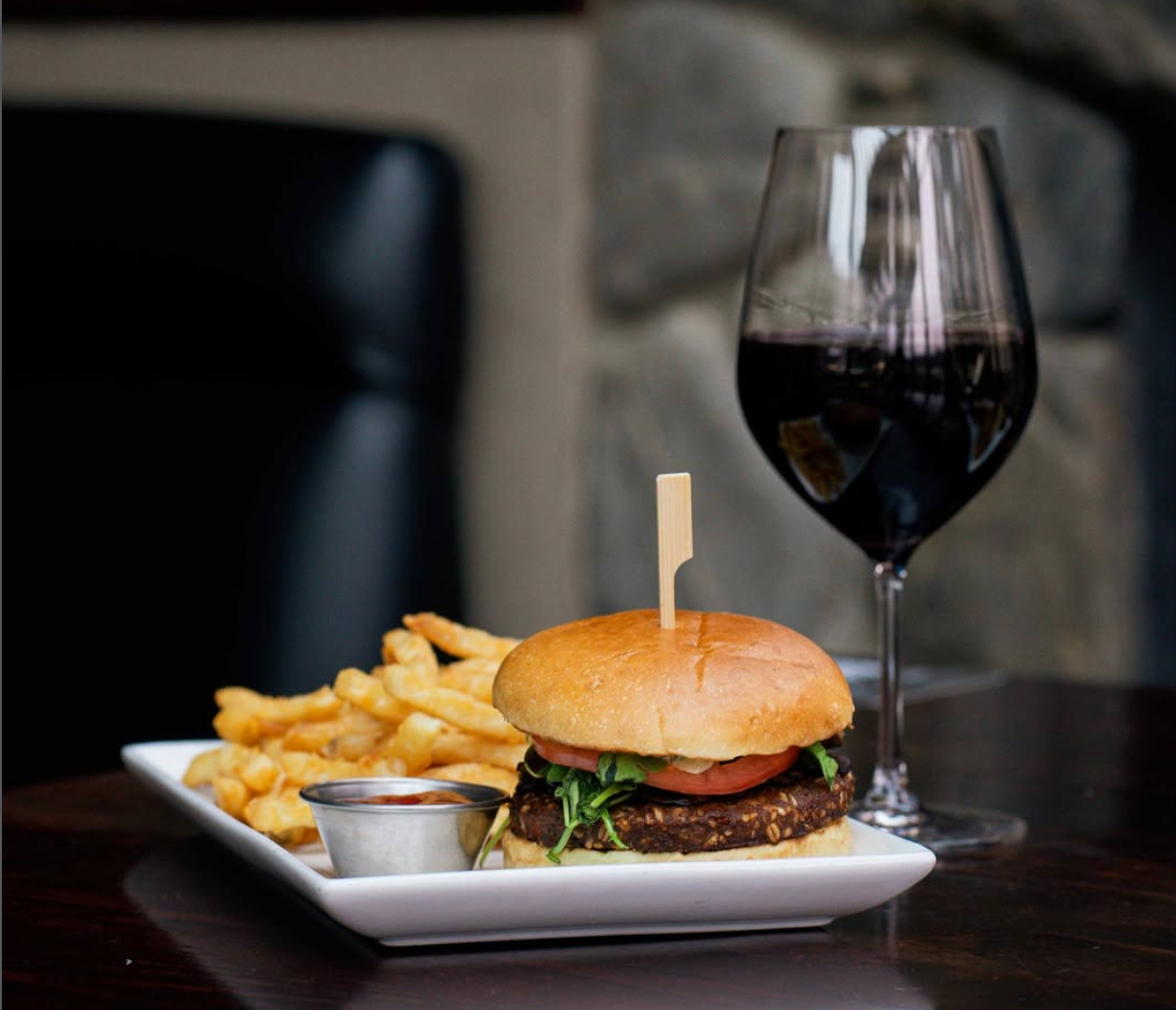 Gourmet burger with fresh greens and tomato served alongside crispy fries and dipping sauce, paired with a glass of red wine on a dark wooden table.