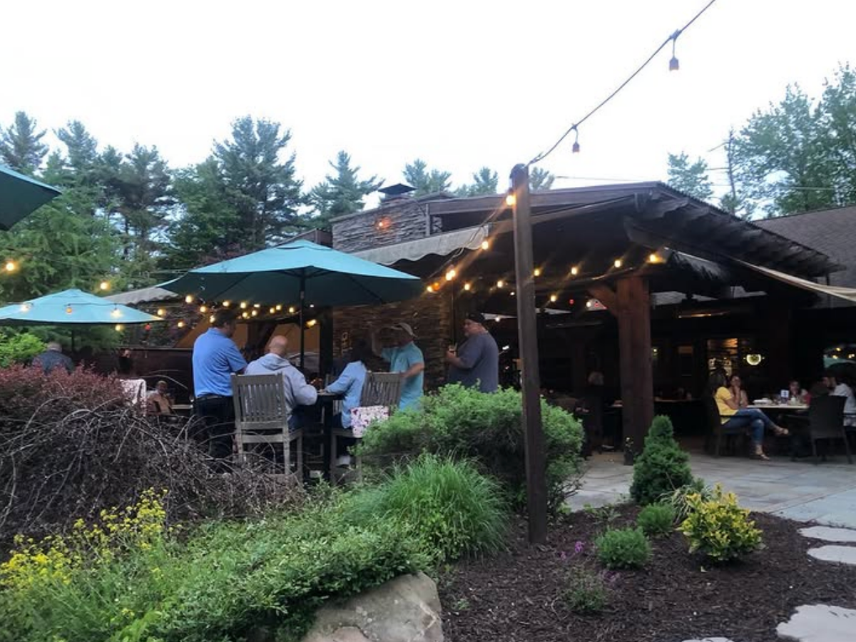 Outdoor restaurant patio with string lights, green umbrellas, and diners enjoying an evening meal surrounded by lush landscaping.