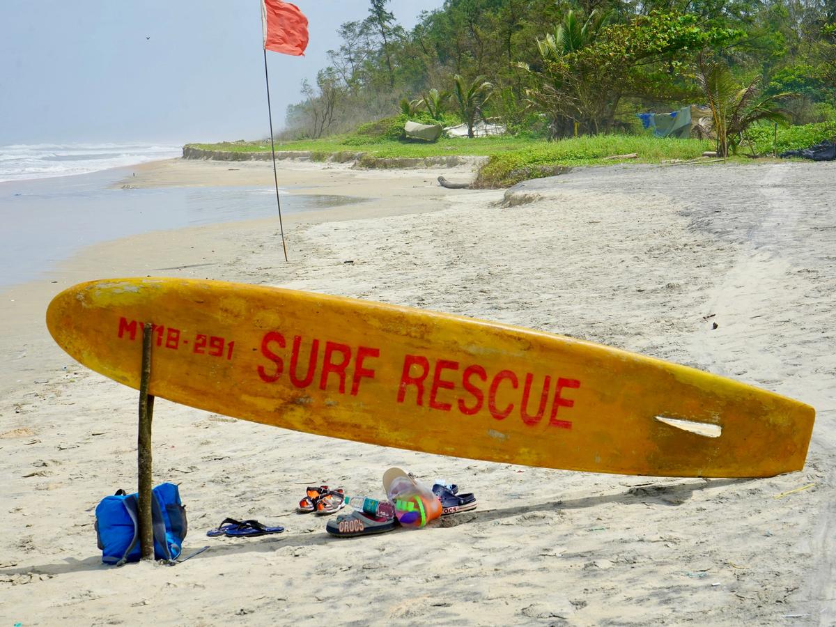 A yellow surf rescue board with red lettering leans against a stick on a sandy beach, next to flip-flops and bags, with a red warning flag and palm trees in the background.
