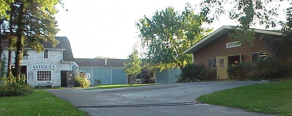 A rustic cluster of antique shop buildings sits along a paved driveway framed by lush green trees, including a white stone barn with an "Antiques" sign and a wood-sided shop with a matching sign. Late-day sunlight filters through the leafy surroundings on a quiet, inviting day.