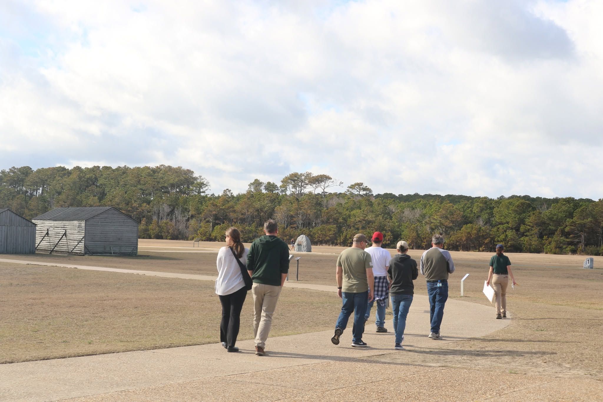 A group of visitors follows a park guide along a paved path across the open grounds of the Wright Brothers National Memorial, with a reconstructed wooden hangar and camp building on the left. A tree line stretches across the horizon beneath a broad, cloud-filled sky.
