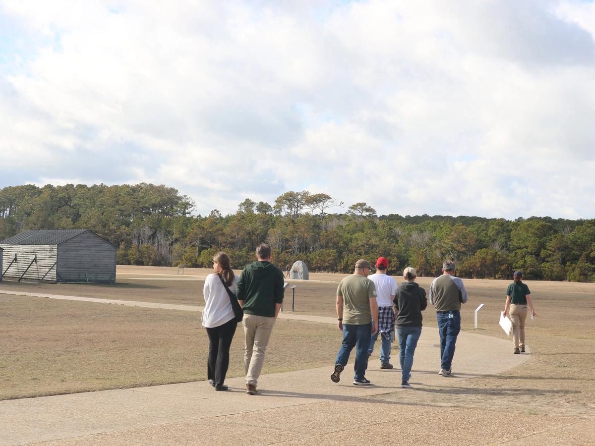 A group of visitors follows a park guide along a paved path across the open grounds of the Wright Brothers National Memorial, with a reconstructed wooden hangar and camp building on the left. A tree line stretches across the horizon beneath a broad, cloud-filled sky.
