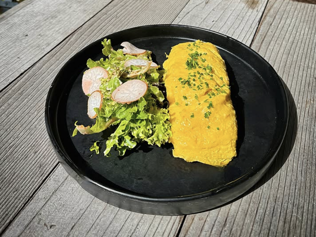 A bright yellow omelette topped with fresh herbs, served with a side salad of frisée lettuce and thinly sliced radishes, all presented on a matte black plate over a rustic wooden surface.