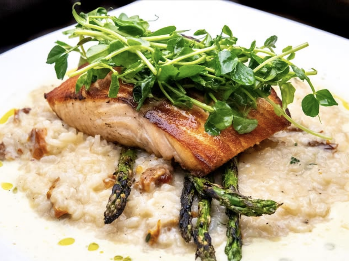 Seared salmon filet topped with fresh greens, served over creamy risotto with grilled asparagus on a white plate.
